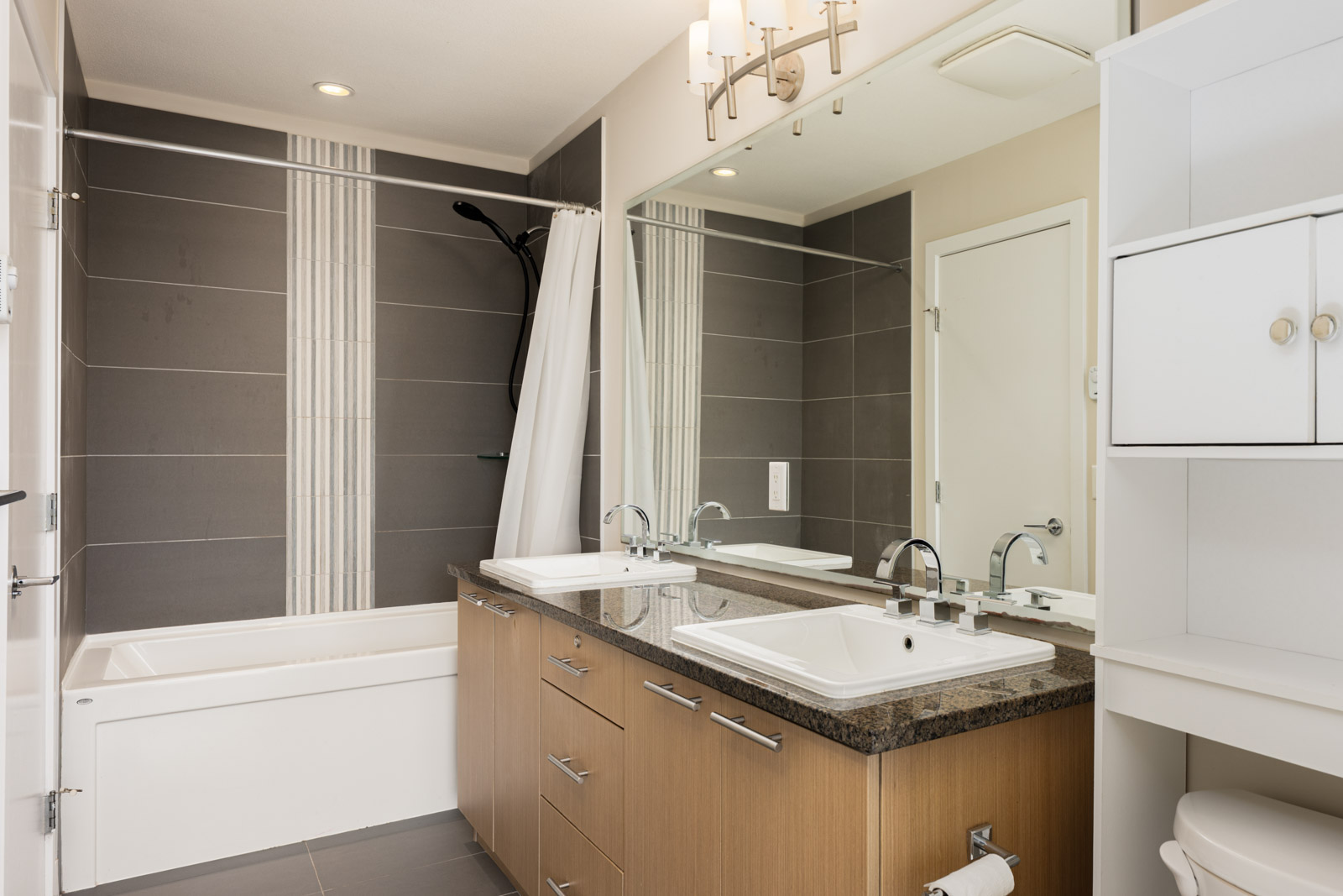 Modern bathroom with double sinks, large mirror, a granite countertop, wood cabinets, built-in shelving, and a bathtub with a shower and white curtain.