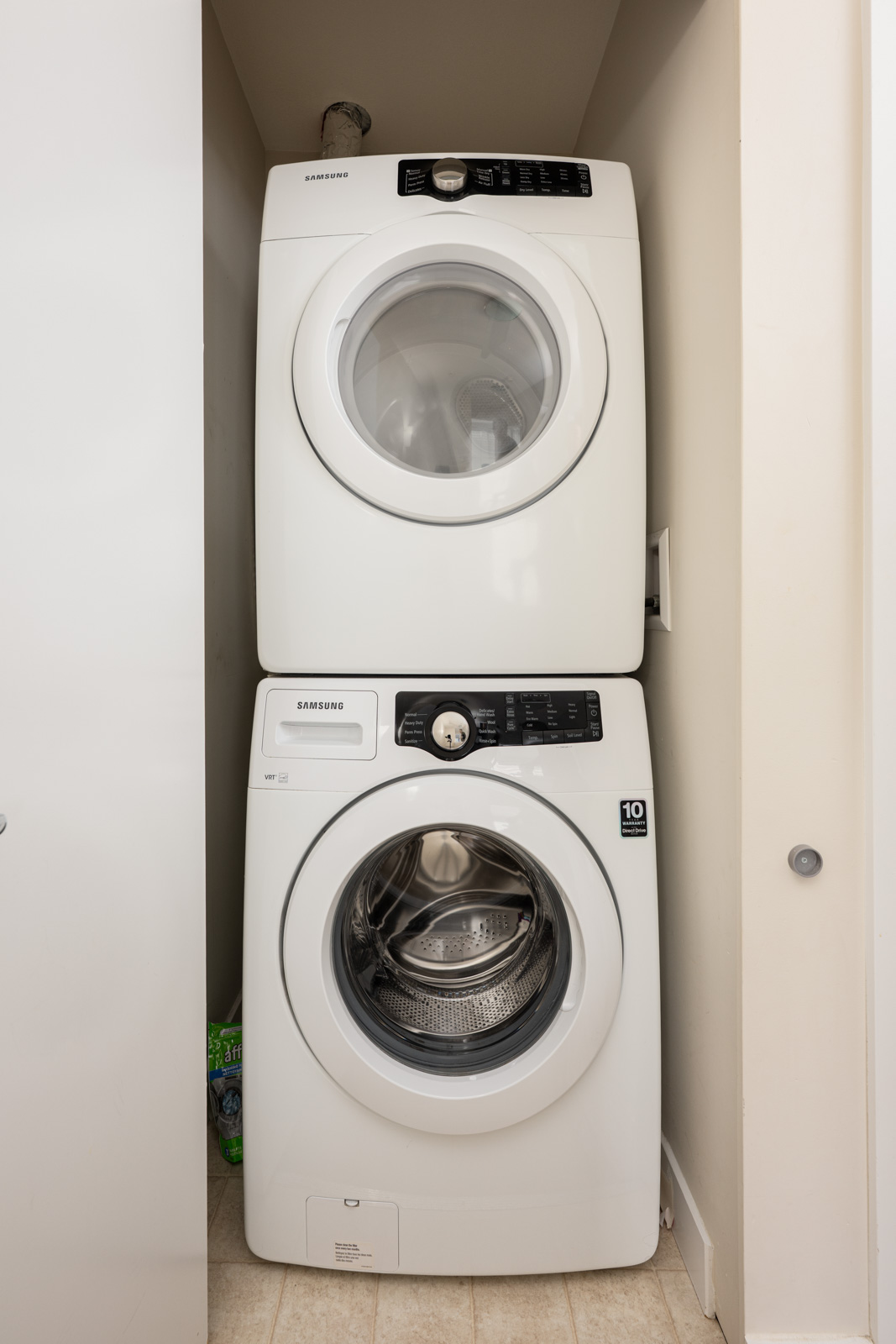A stacked Samsung washer and dryer set in a narrow laundry closet with beige walls and tiled flooring.