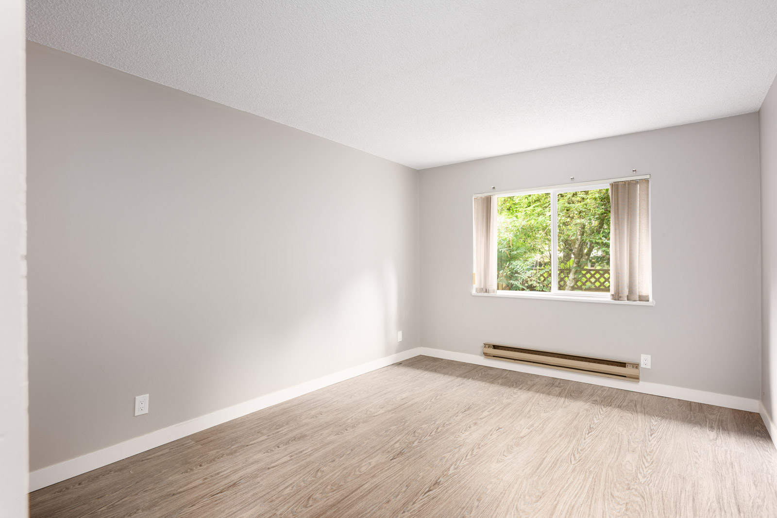 Empty room with light gray walls, wood-look flooring, a large window with vertical blinds, and a baseboard heater below the window.