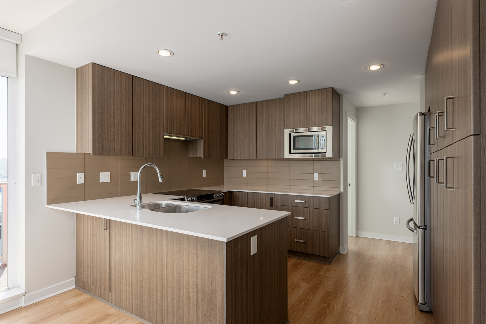 Modern kitchen with wood cabinets, white countertops, built-in microwave, stainless steel appliances, and light wood flooring.