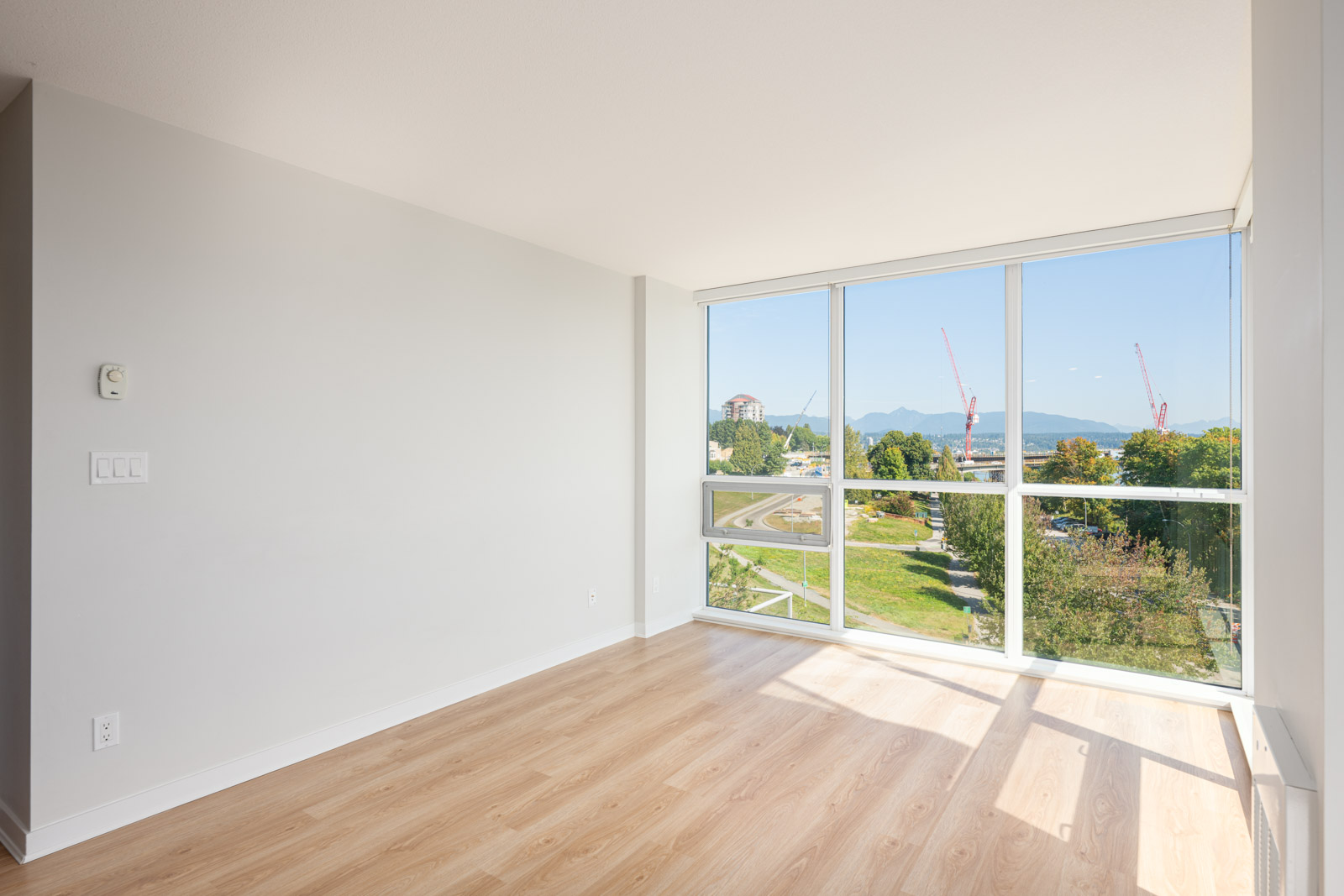 Bright empty room with light wood floors, white walls, and large windows offering a view of trees, buildings, and distant mountains under a clear blue sky.
