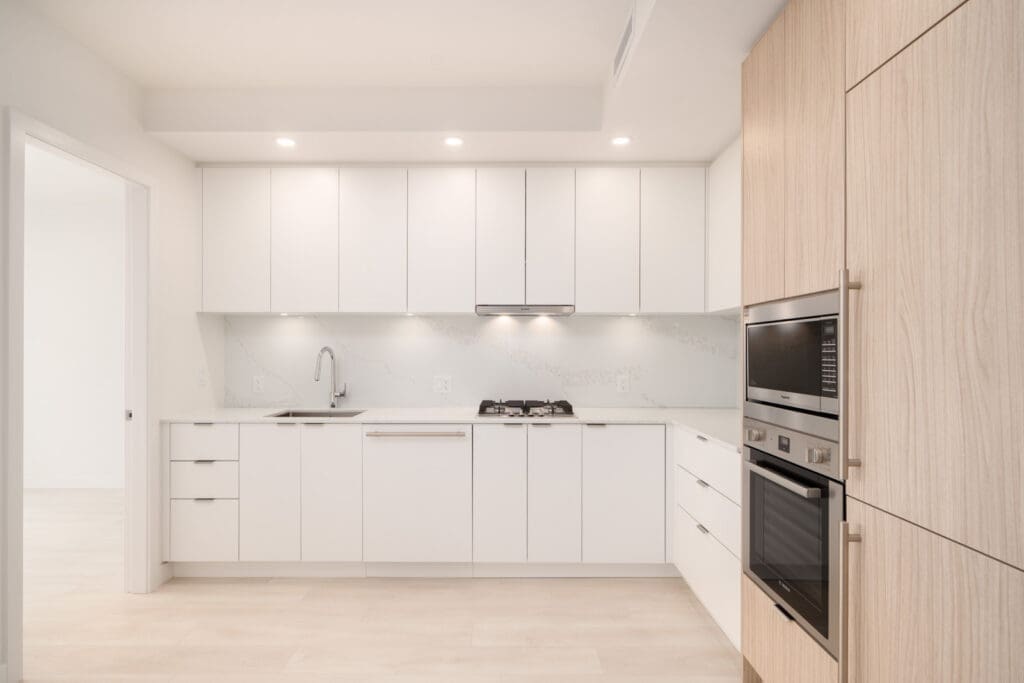 Modern kitchen with white cabinets, light wood accents, built-in oven and microwave, gas cooktop, and under-cabinet lighting.