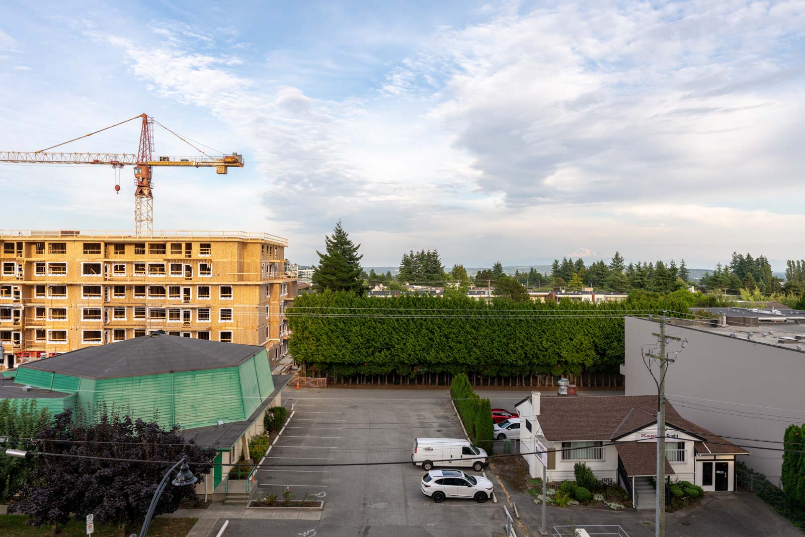 A construction site with a crane next to an unfinished building, adjacent to a parking lot with cars, trees, and several low-rise buildings under a partly cloudy sky.