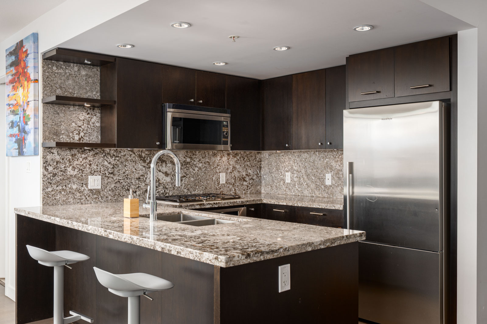 Modern kitchen with dark wood cabinets, stainless steel appliances, granite countertops, a double sink, and two white bar stools at the island.