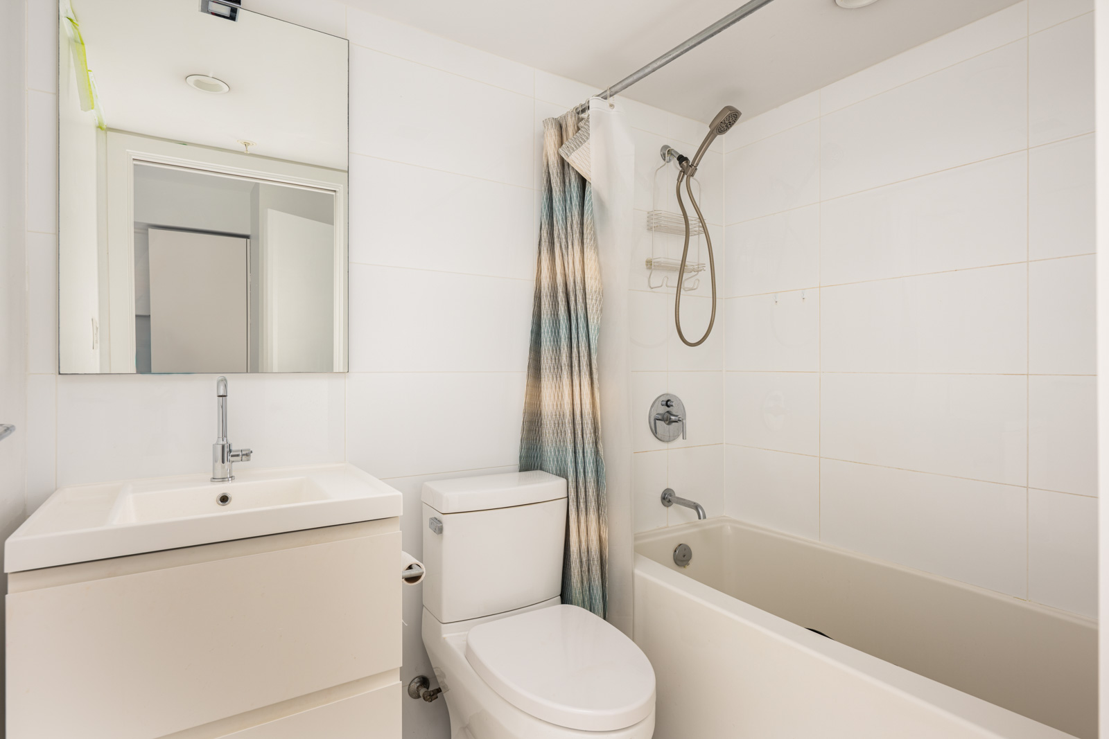 Modern bathroom with a white sink, toilet, and bathtub. A striped shower curtain is partially drawn, and a wall mirror is above the sink. The walls and tiles are white.