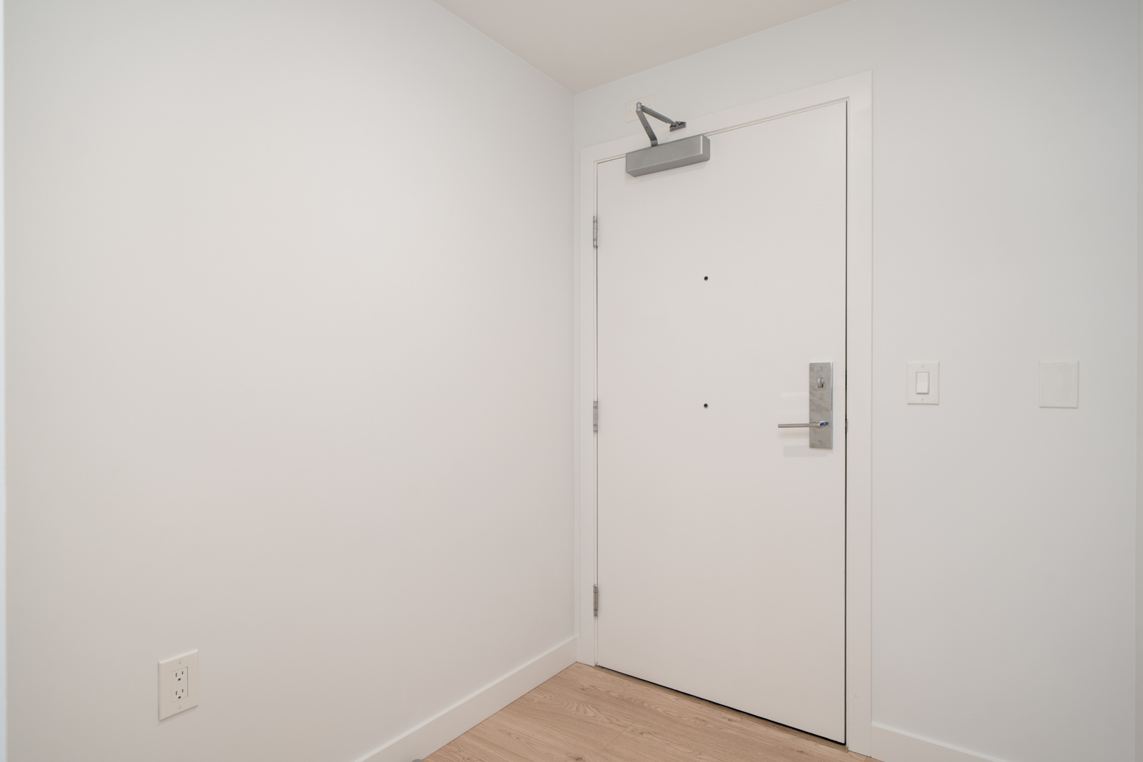 A plain white entry door with a metallic handle, door closer, and keypad, set in a corner of a minimalist room with white walls and light wood flooring.