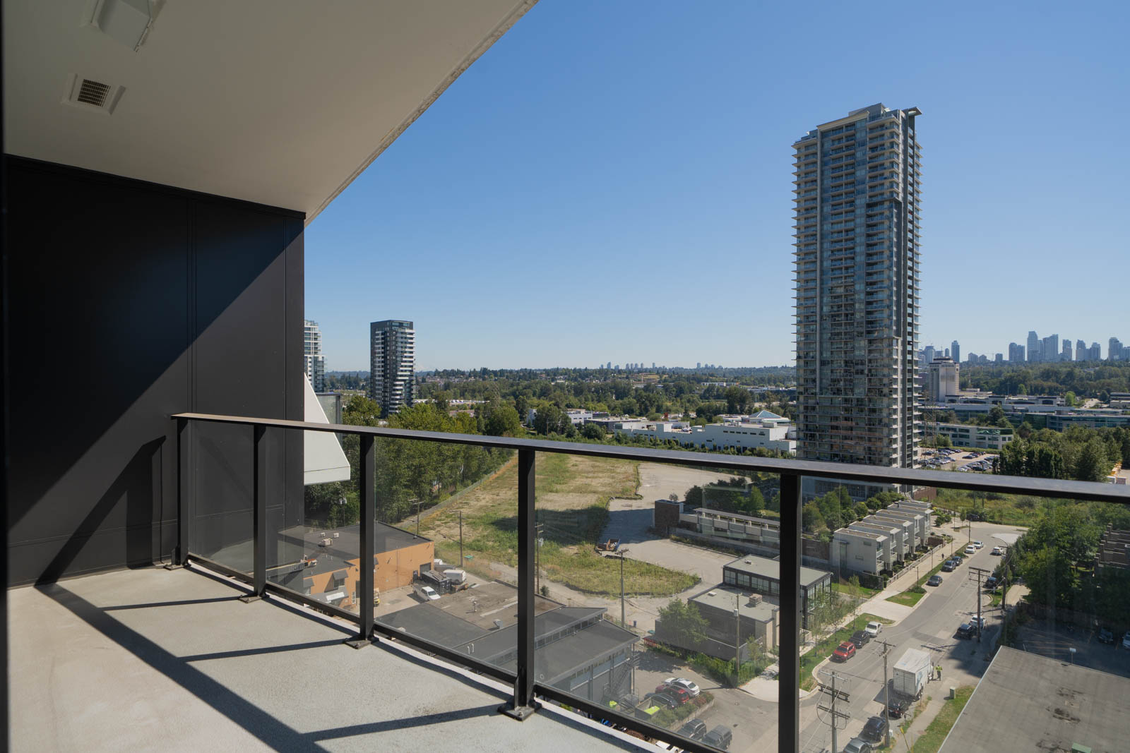 A modern balcony with glass railing overlooks a cityscape featuring high-rise buildings, trees, and clear blue sky.