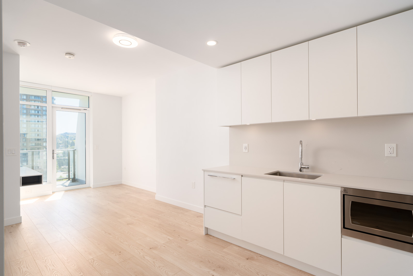 Modern, unfurnished apartment kitchen with white cabinets, light wood flooring, built-in appliances, and large windows leading to a balcony with a city view.