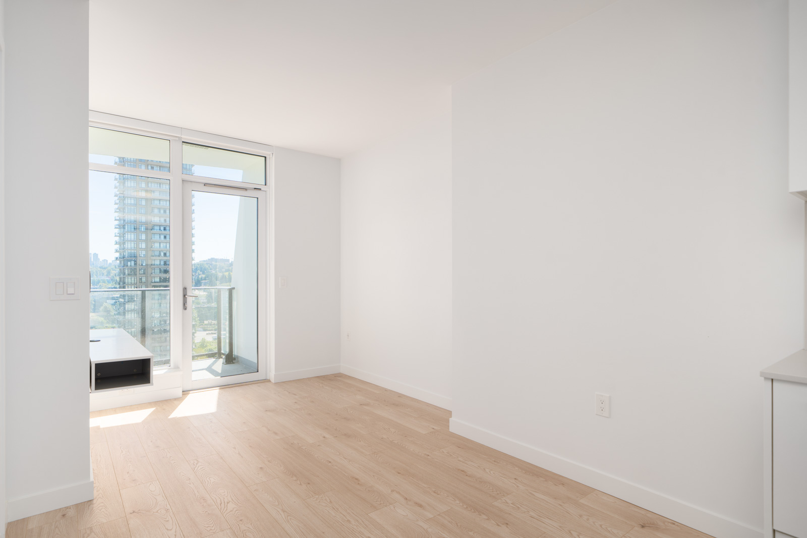 A bright, empty apartment room with light wood flooring, white walls, large windows, and a balcony overlooking a cityscape.