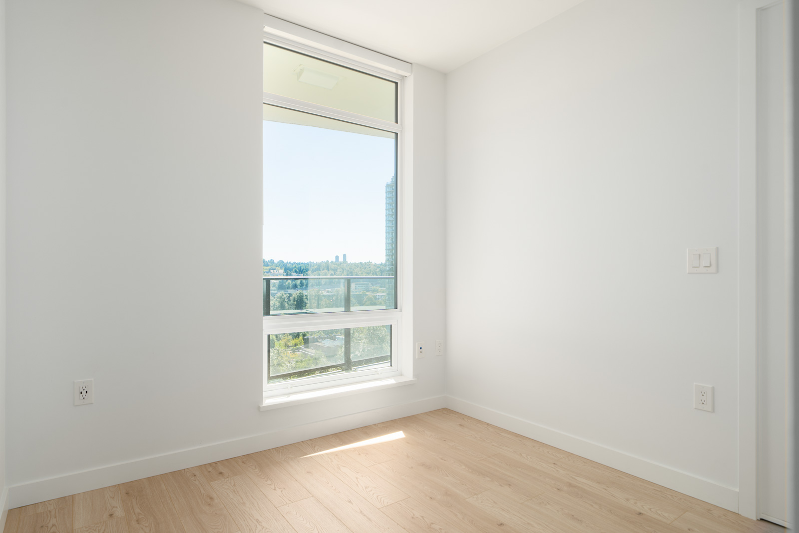 Empty room with light wood flooring, white walls, and a large window letting in natural light with a view of buildings and greenery outside.