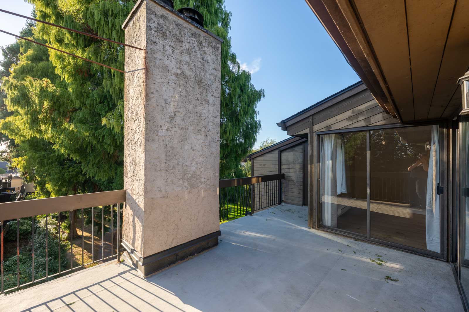 A concrete balcony with a stucco chimney, metal railing, and sliding glass doors leading inside; trees and greenery visible in the background.