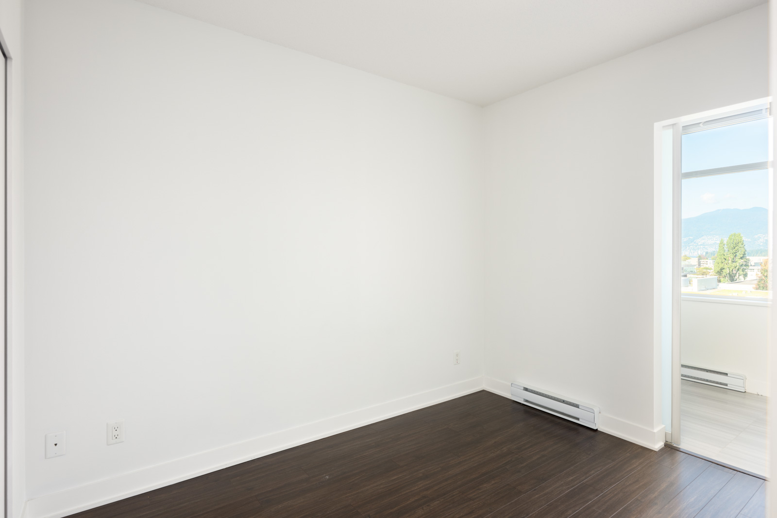 Empty room with white walls, dark wood floor, baseboard heater, electrical outlets, and a window allowing natural light and an outdoor view.