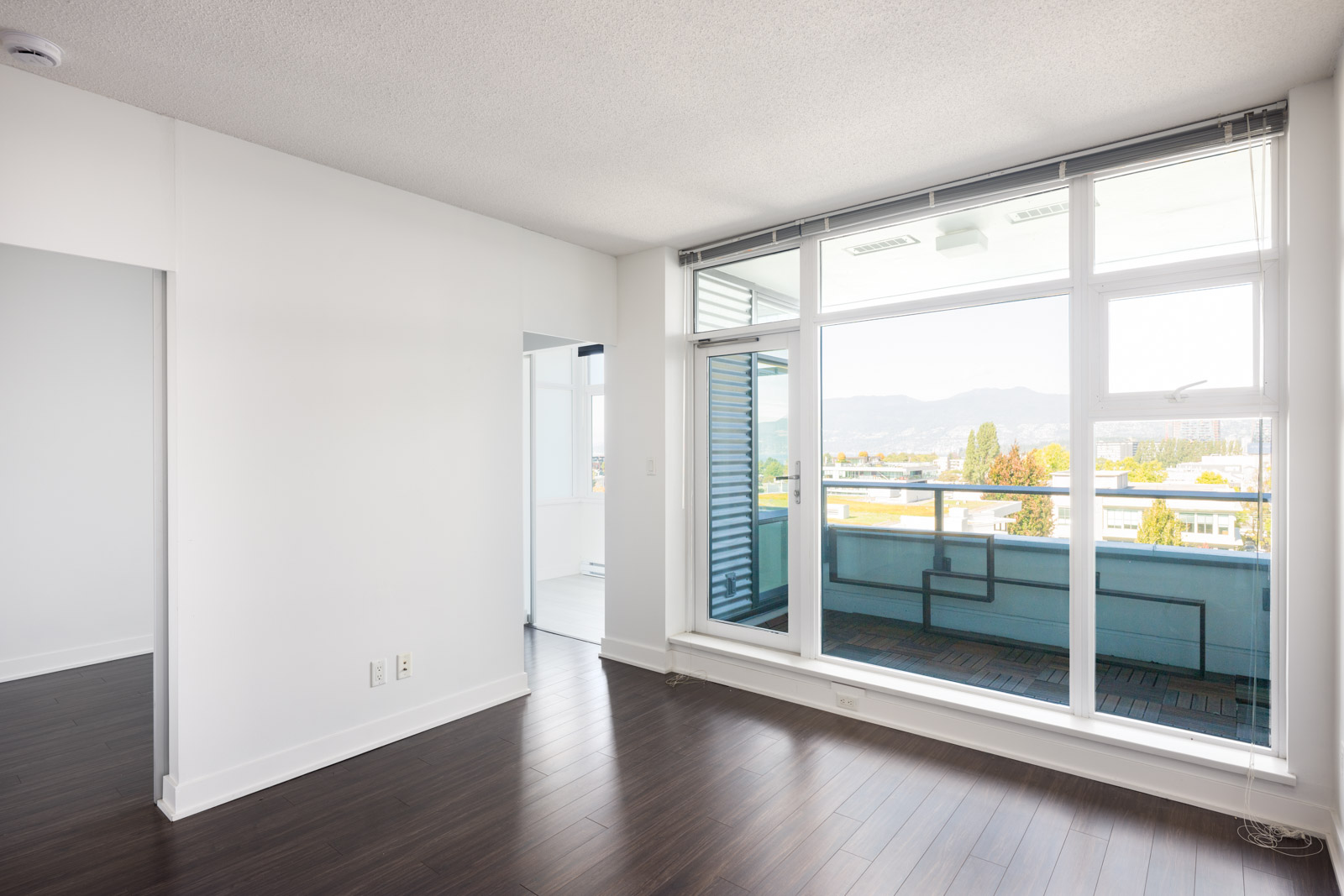 Bright empty room with dark wood floors, white walls, large windows, and a glass door leading to a balcony overlooking a cityscape and distant mountains.