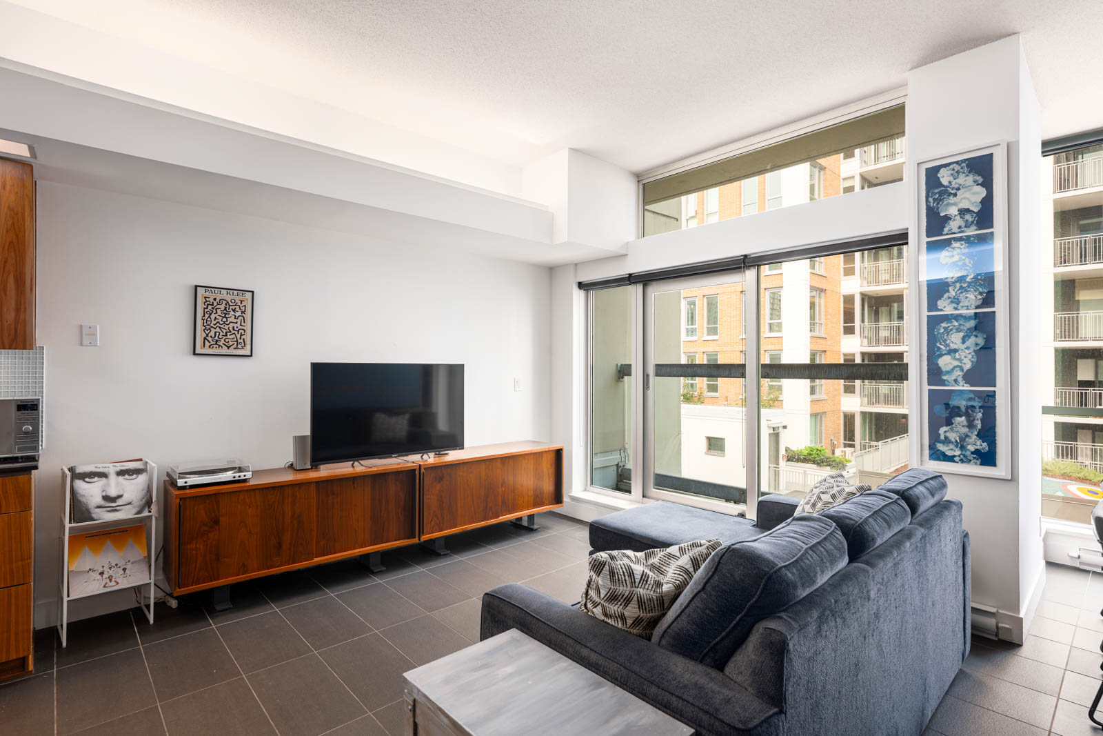 Modern living room with a blue sofa, wooden TV console, flat-screen TV, wall art, and large windows overlooking apartment buildings.