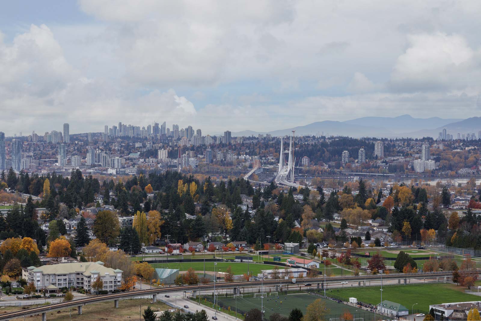 A cityscape view showing urban buildings, bridges, and residential neighborhoods surrounded by autumn trees with mountains in the background under a partly cloudy sky.