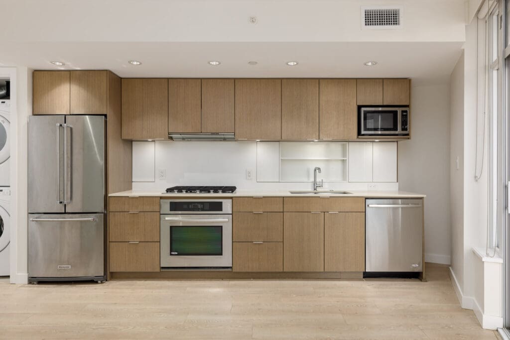 Modern kitchen with light wood cabinets, stainless steel appliances, gas stove, built-in oven, microwave, dishwasher, and a stacked washer and dryer unit on the left.