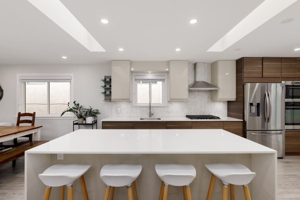 Modern kitchen with white island, three white barstools, stainless steel appliances, wooden cabinets, and a dining table with chairs on the left side.