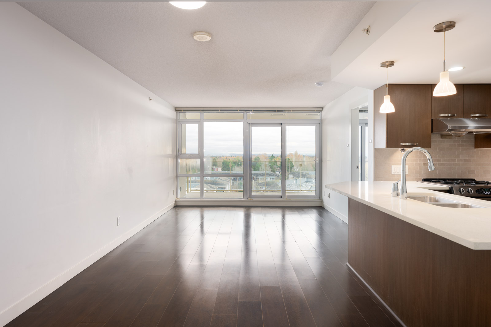 Unfurnished modern apartment with dark wood floors, white walls, large windows, and an open kitchen area with pendant lights and stainless steel appliances.