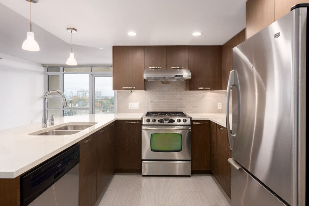 Modern kitchen with stainless steel appliances, brown cabinets, white countertops, and overhead lighting, featuring a gas stove, dishwasher, and large refrigerator.