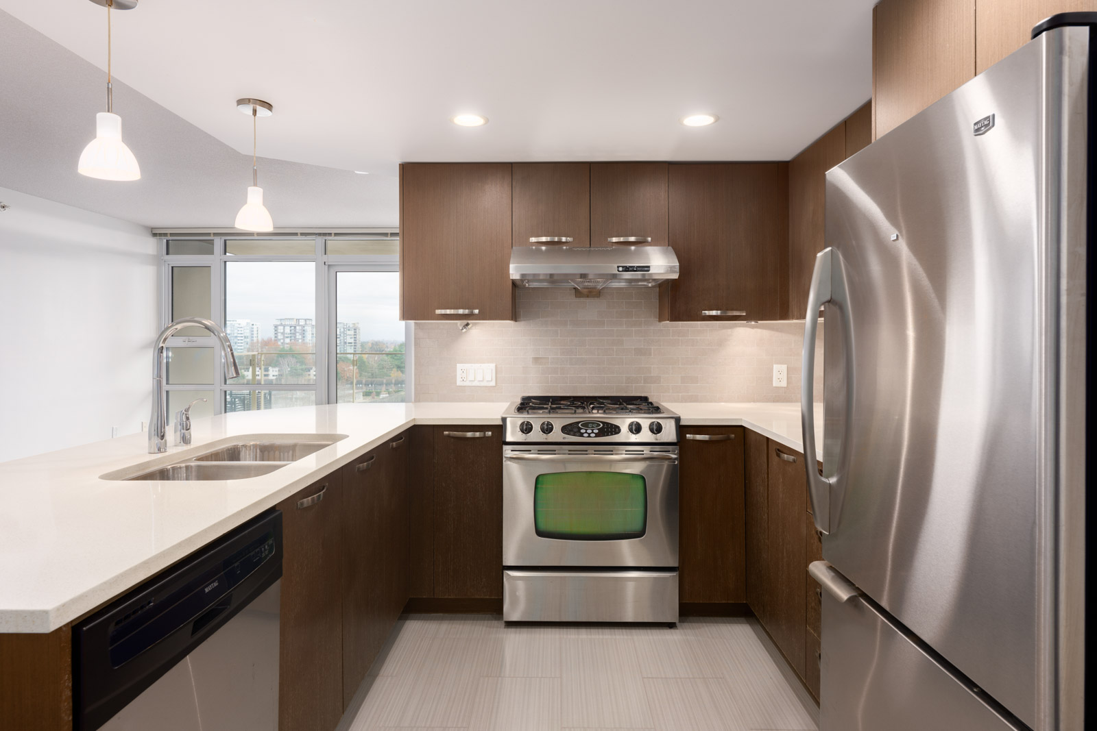 Modern kitchen with stainless steel appliances, brown cabinets, white countertops, and overhead lighting, featuring a gas stove, dishwasher, and large refrigerator.