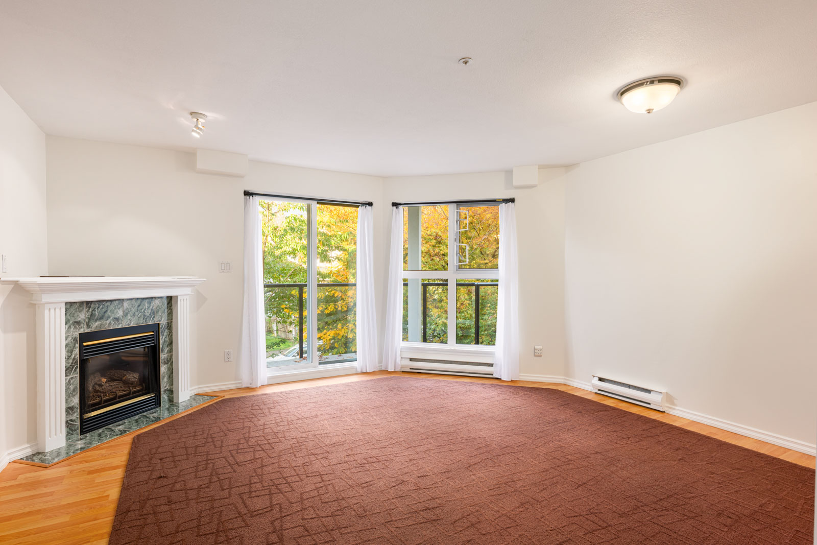 Empty living room with a fireplace, large bay windows with white curtains, brown carpet, and white walls. Trees with autumn leaves are visible outside the windows.