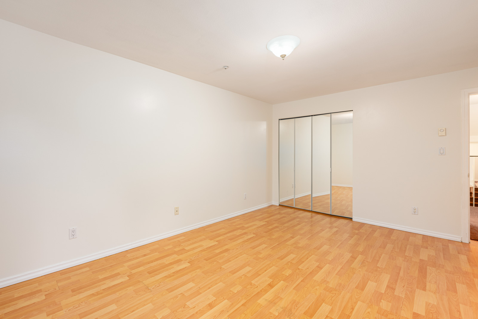 Empty room with light-colored walls, wood laminate flooring, ceiling light fixture, and a closet with mirrored sliding doors.