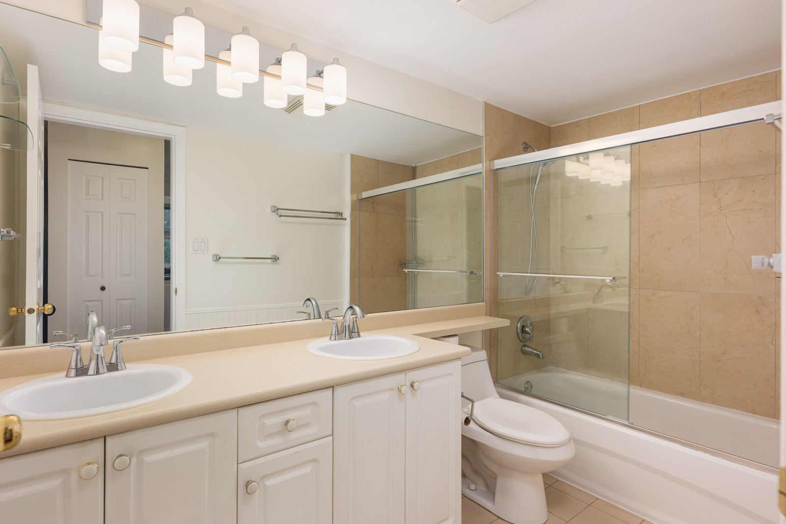 Modern bathroom with double sink vanity, large mirror, white cabinets, toilet, and a bathtub with glass sliding doors and beige tile walls.