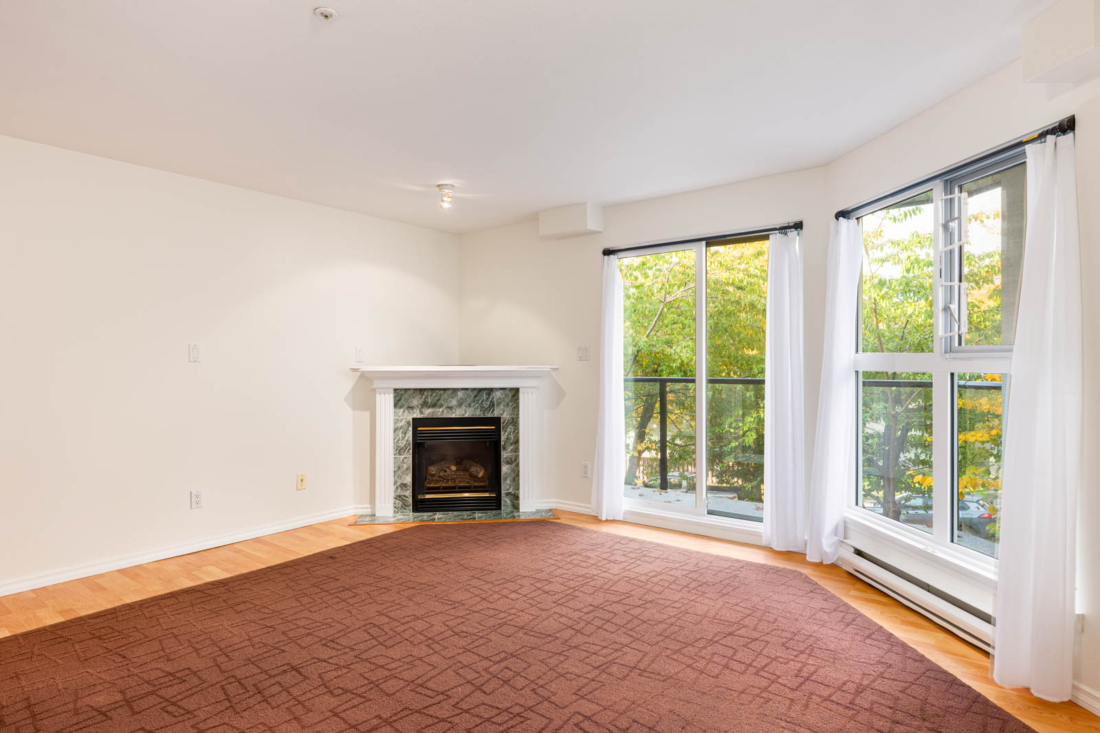 Unfurnished living room with a brown carpet, corner fireplace, large windows with white curtains, and a view of trees outside.