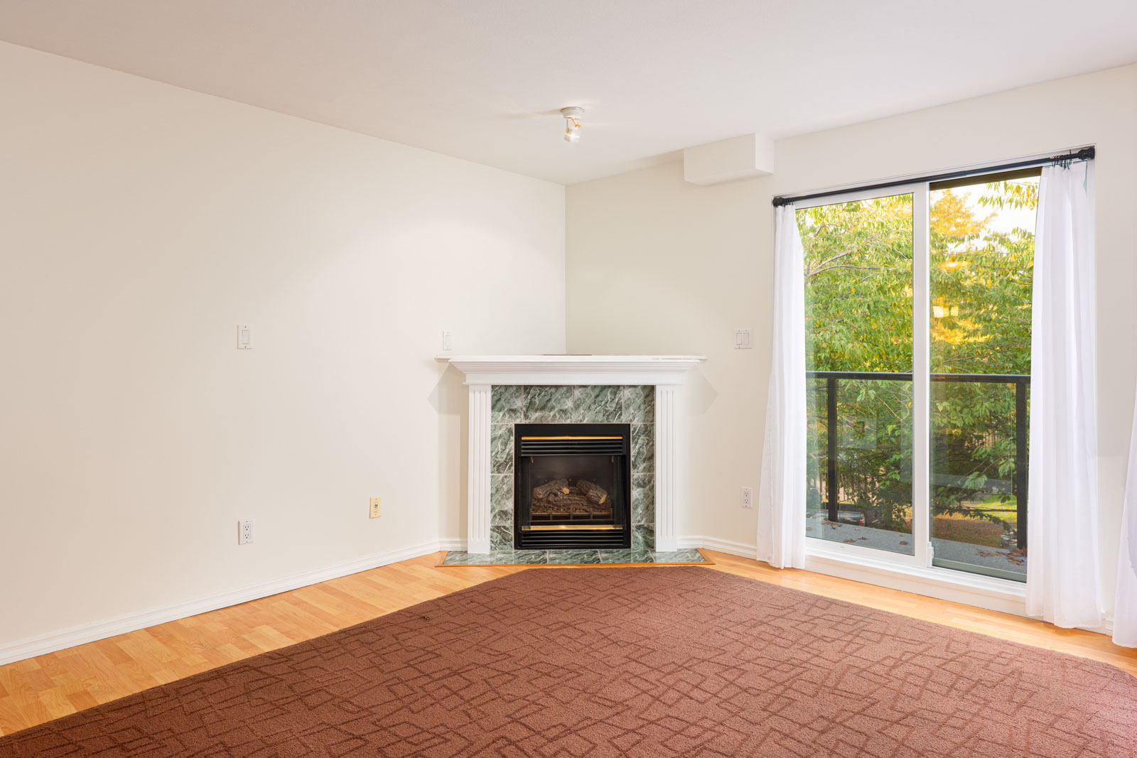 Unfurnished living room with a fireplace, a large window with white curtains, light-colored walls, and hardwood flooring with a brown carpet.