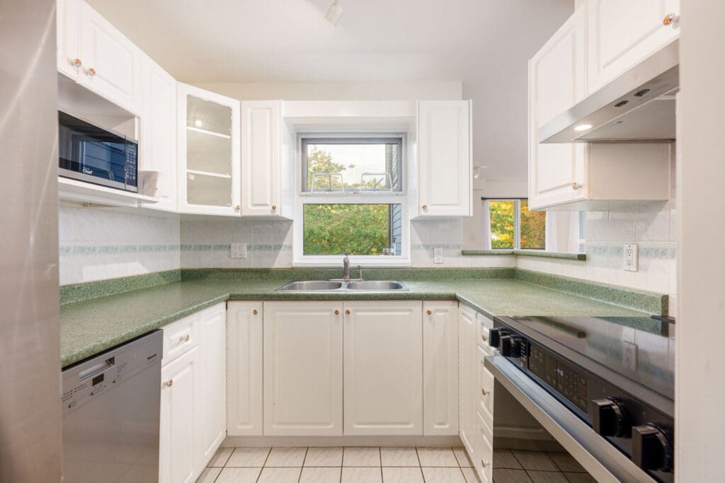 A small kitchen with white cabinets, green countertops, a double sink under a window, and stainless steel appliances.