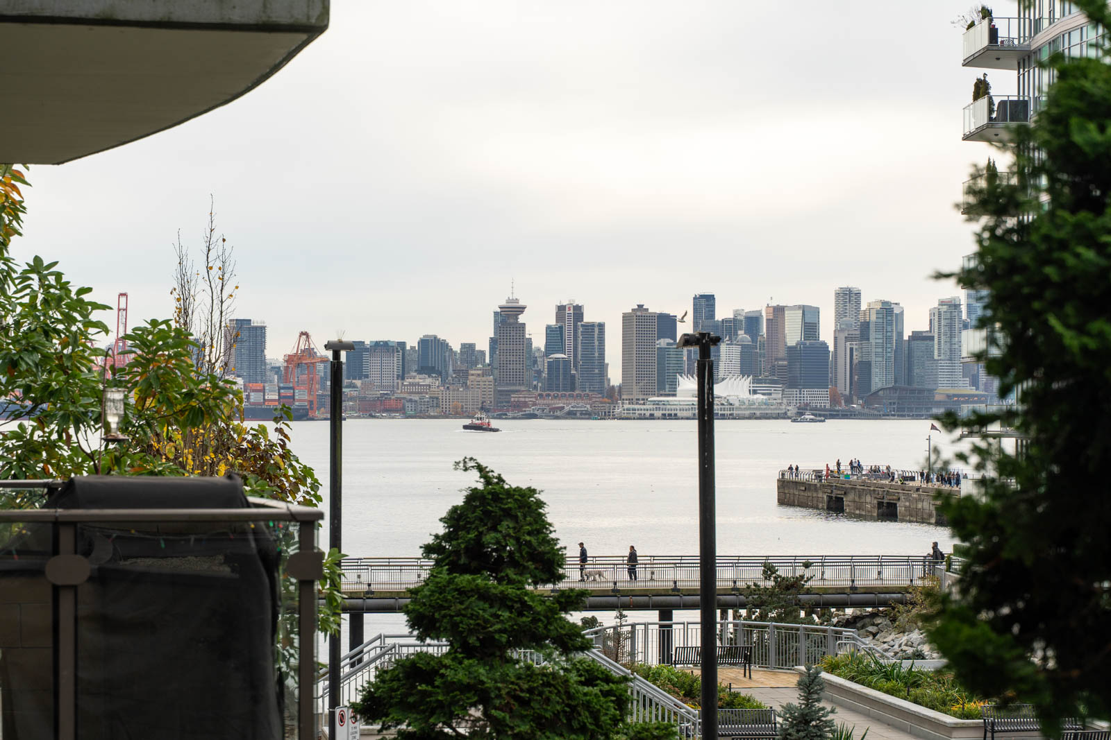 View of a city skyline across a body of water, with people walking along a pier in the foreground and buildings and trees framing the scene.