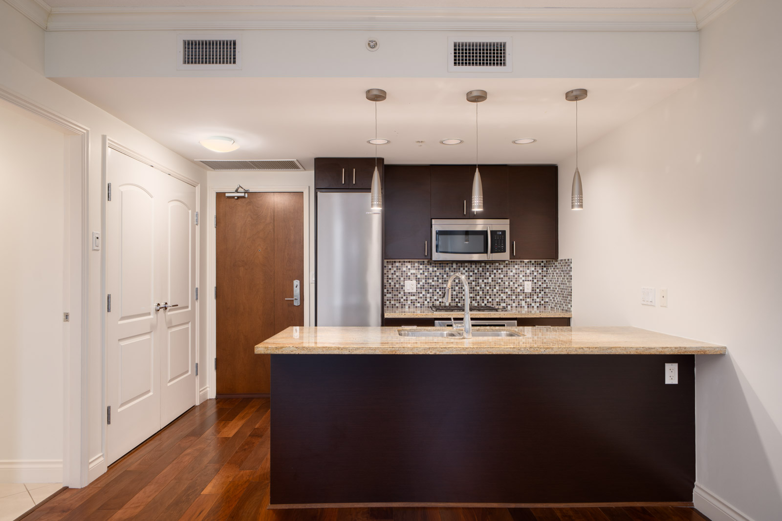 Modern kitchen with dark wood cabinets, stainless steel appliances, mosaic tile backsplash, and a central island with a sink and pendant lighting.