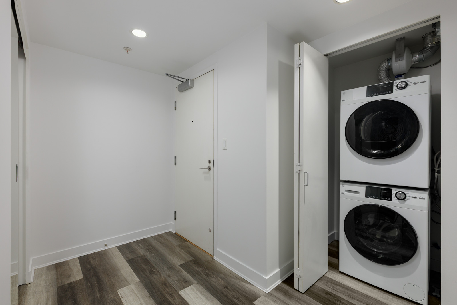 A small laundry area with a stacked washer and dryer behind folding doors next to a white entrance door in a modern room with wood-patterned flooring.
