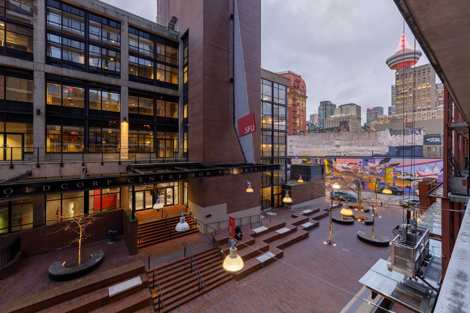 A modern urban courtyard with brick pathways, outdoor seating, and SFU Goldcorp Centre for the Arts building; city skyscrapers and the Vancouver Lookout tower in the background.
