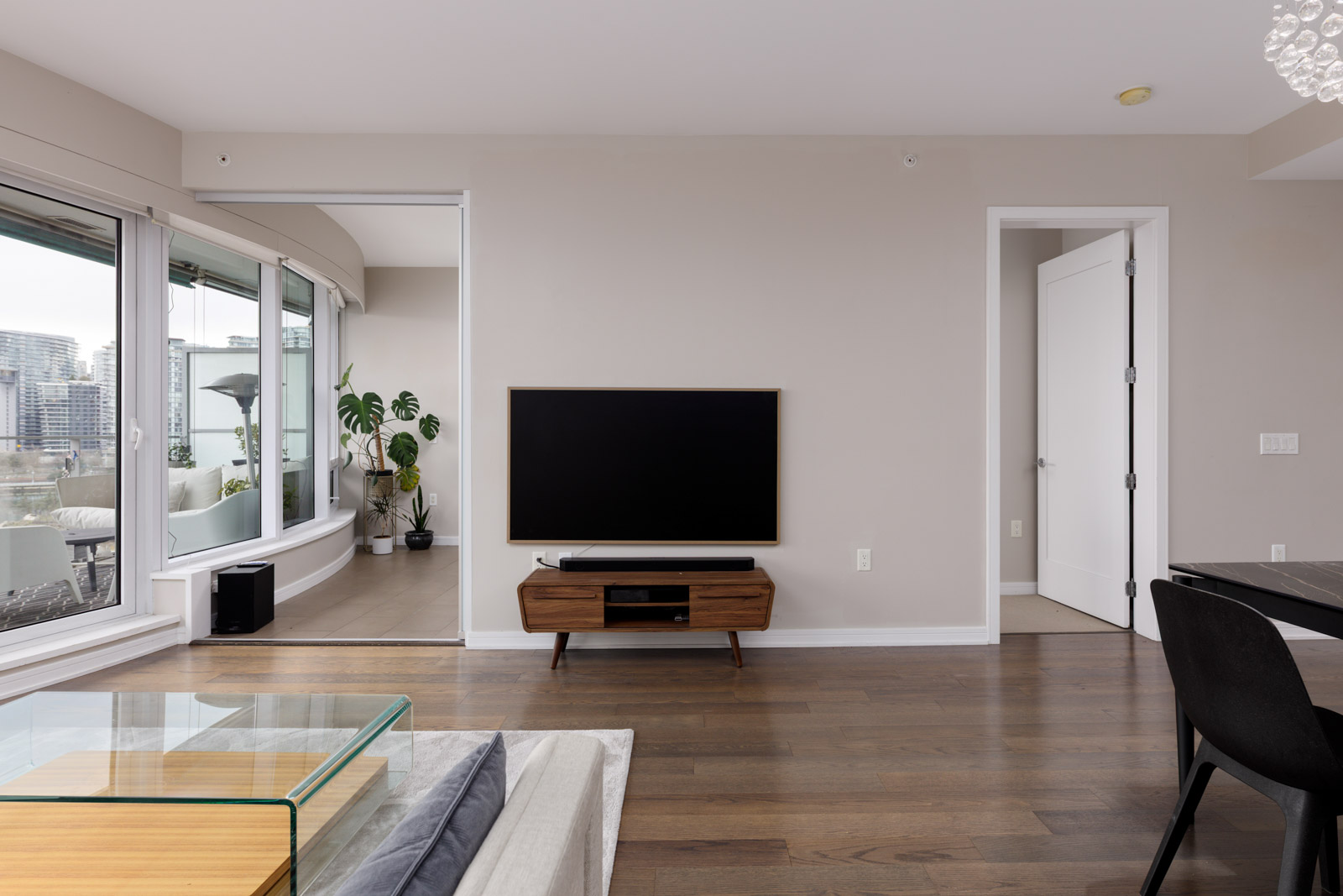 A modern living room with a wall-mounted TV, wooden TV stand, glass coffee table, and large windows overlooking a cityscape.