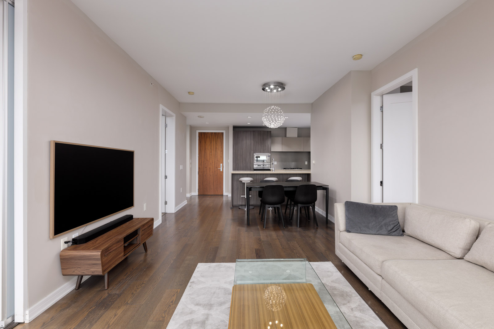 Modern living room with beige sofa, glass coffee table, wall-mounted TV, and open kitchen with dining table and chairs in the background.