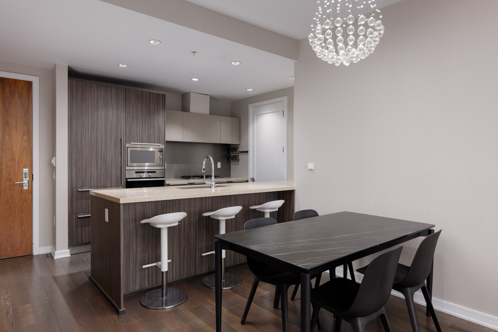 Modern kitchen and dining area with a wooden table, six black chairs, an island with three white stools, stainless steel appliances, and a contemporary light fixture.