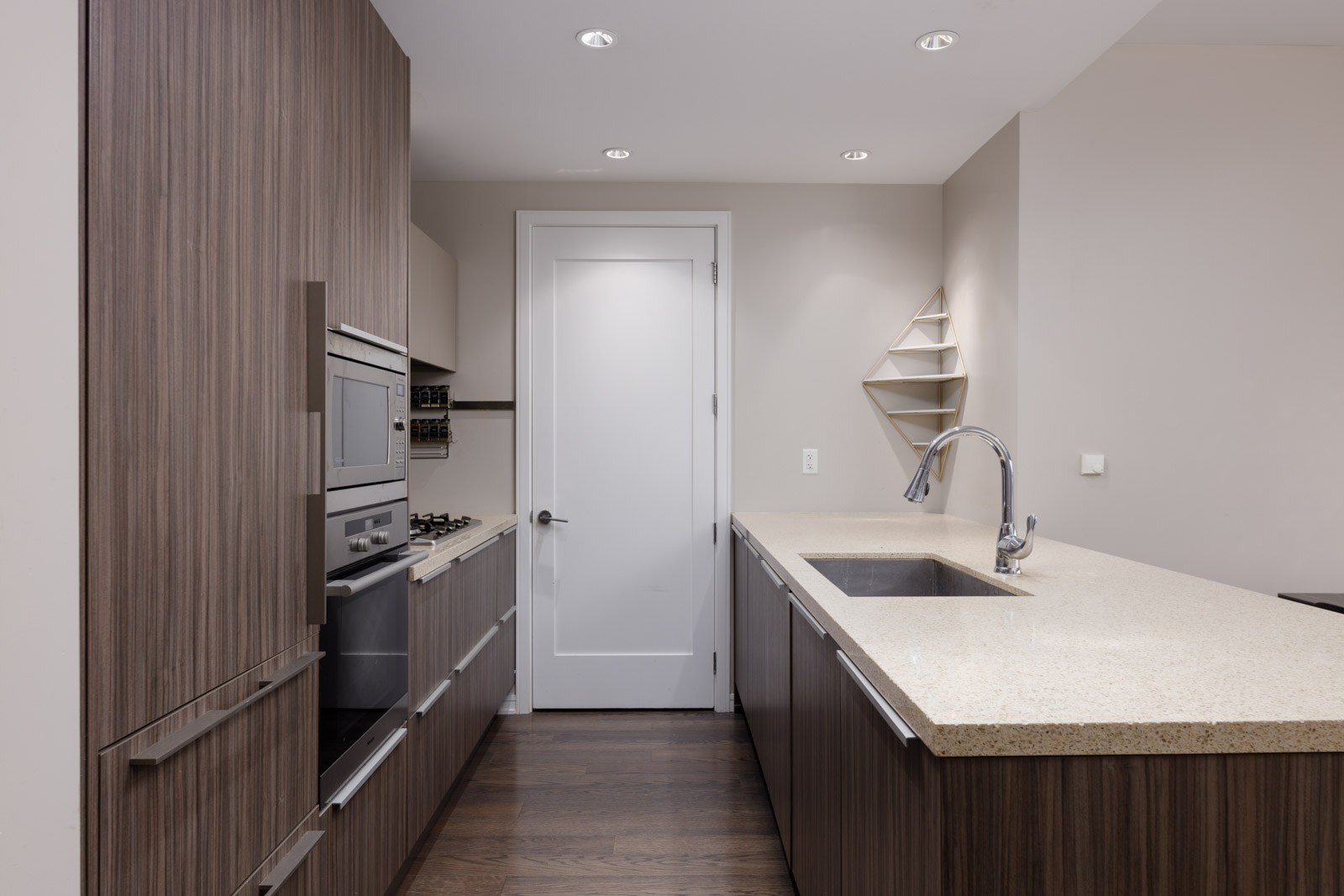 Modern kitchen with wood veneer cabinets, built-in oven, gas stovetop, light countertops, stainless steel faucet, and minimalist decor, featuring a white door and shelf on the back wall.