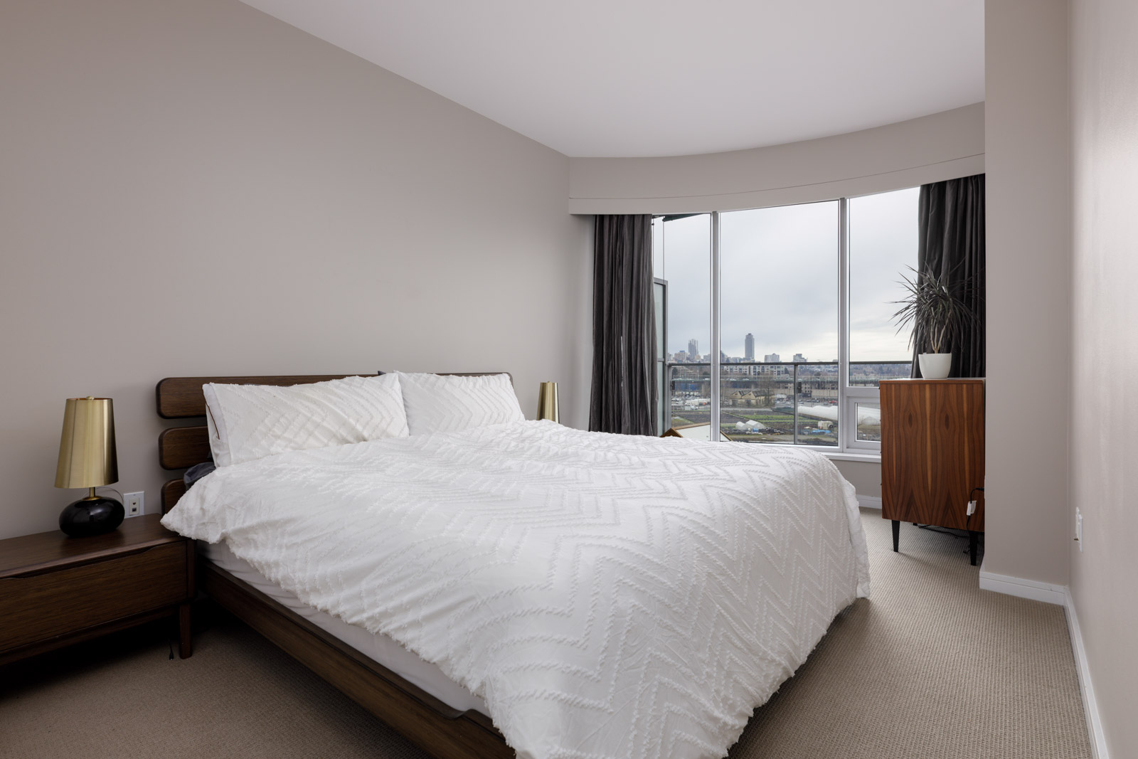 A modern bedroom with a large bed, two side tables with lamps, a dresser, and a window overlooking a cityscape.