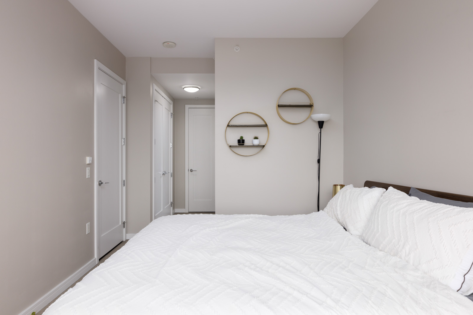A neatly made bed with white bedding in a minimalist bedroom featuring beige walls, round wall shelves, and a floor lamp.