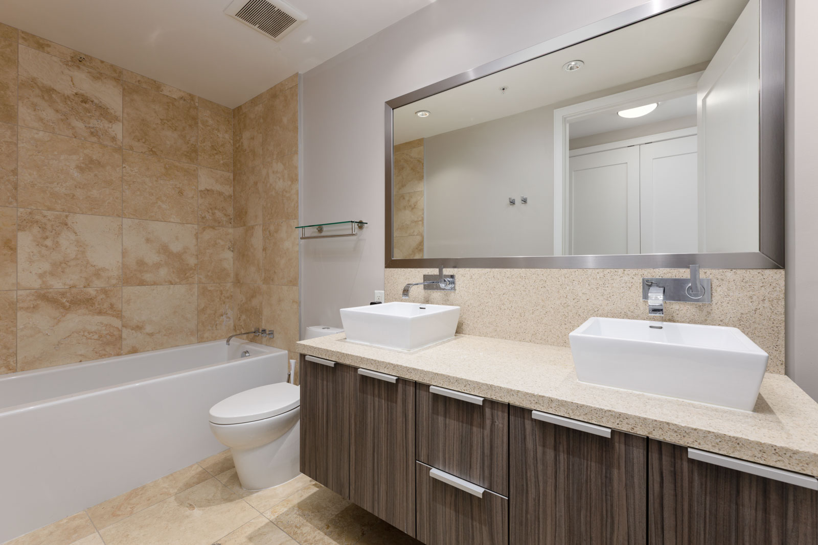 Modern bathroom with beige tile walls, a bathtub, toilet, double sink vanity with vessel sinks, large mirror, and wooden cabinets.
