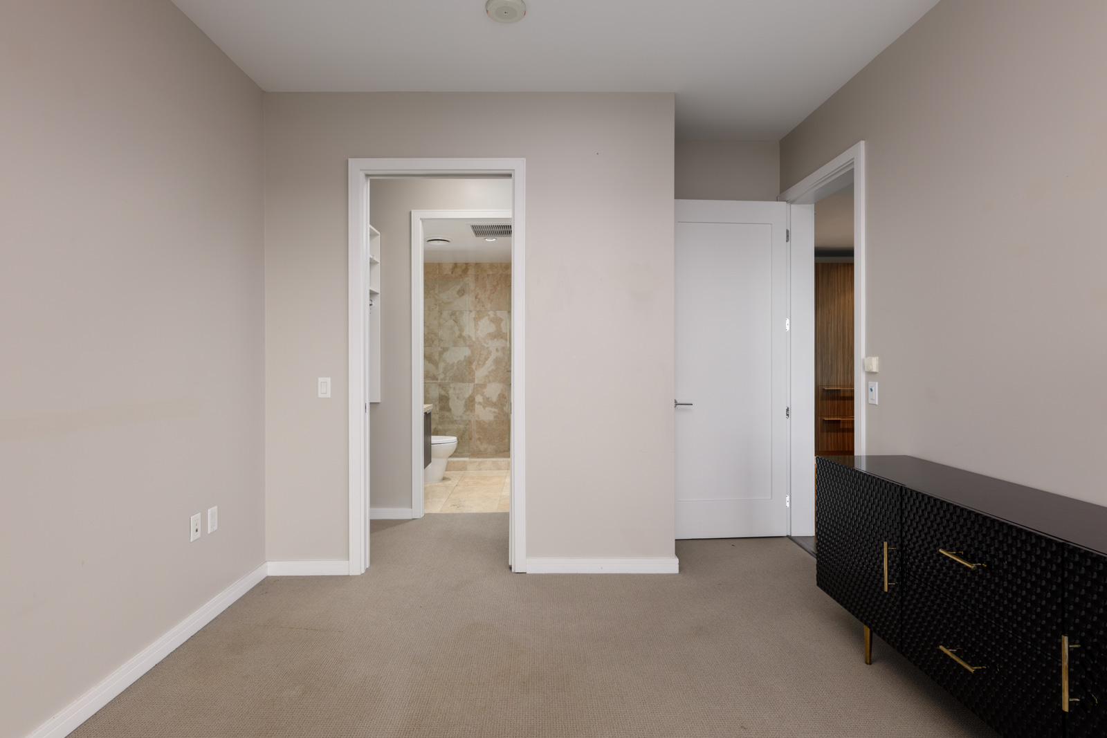 A beige-carpeted room with light-colored walls, a black dresser on the right, and a view into a bathroom with tiled walls through an open doorway.