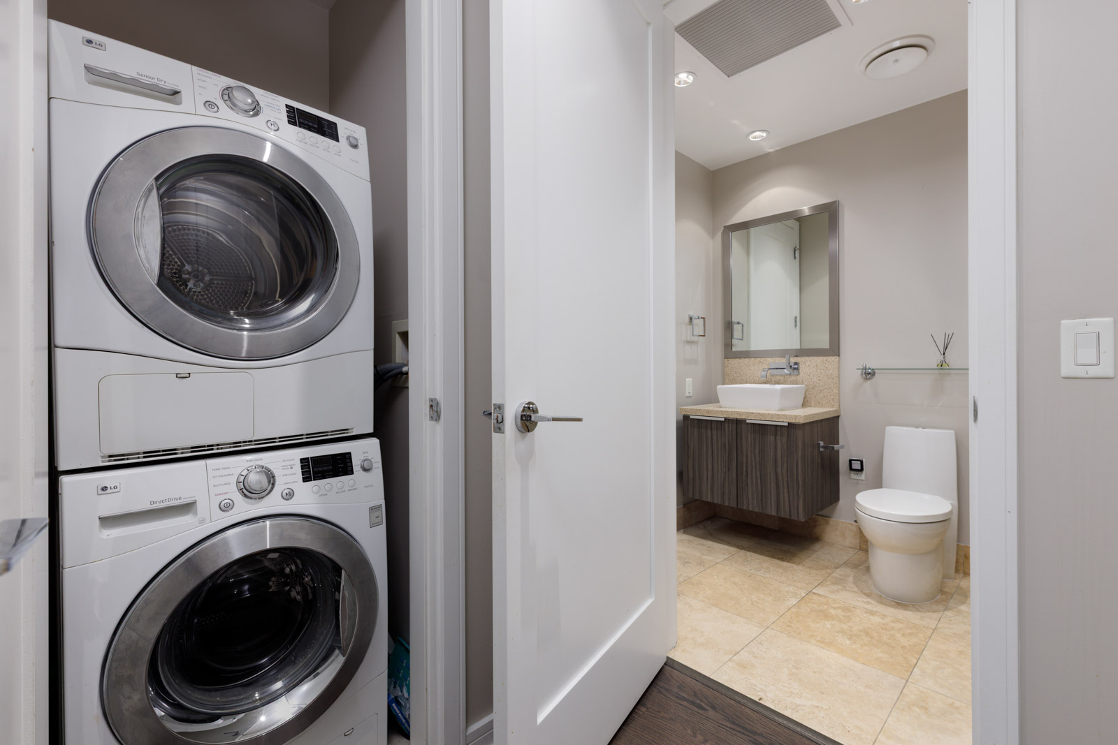 A laundry area with stacked washer and dryer is next to an open door leading to a modern bathroom with a toilet, sink, and mirror.
