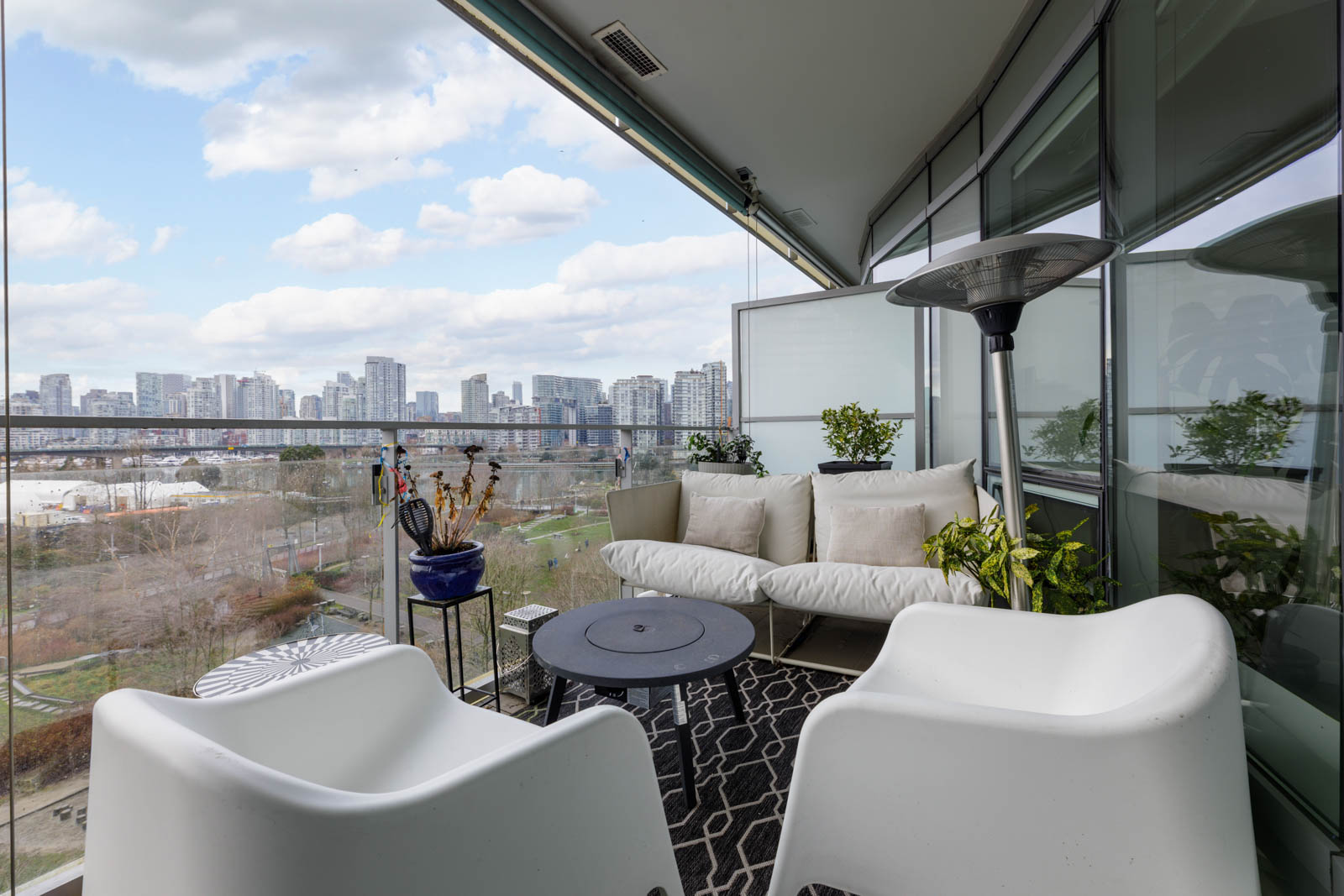 A modern balcony with white chairs, a sofa, potted plants, a patio heater, and a city skyline visible in the background.