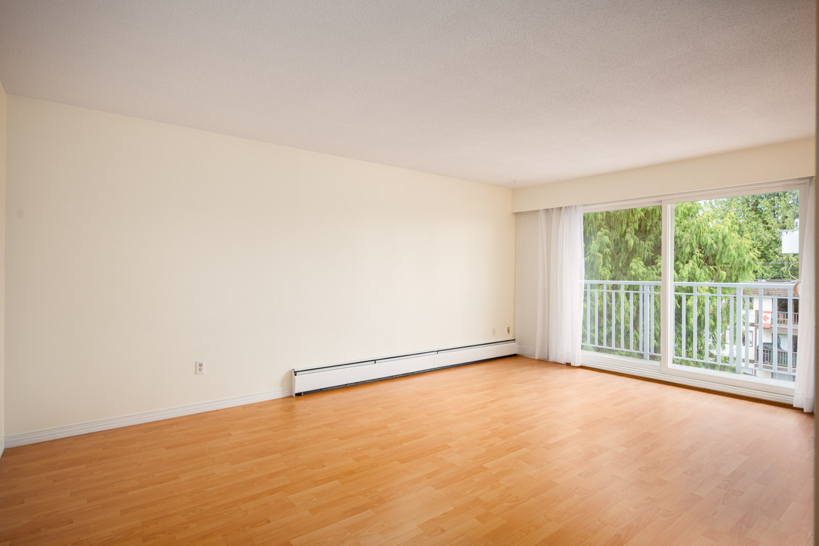 Empty living room with light wood flooring, cream walls, a large window with sliding glass door, and a view of trees outside.