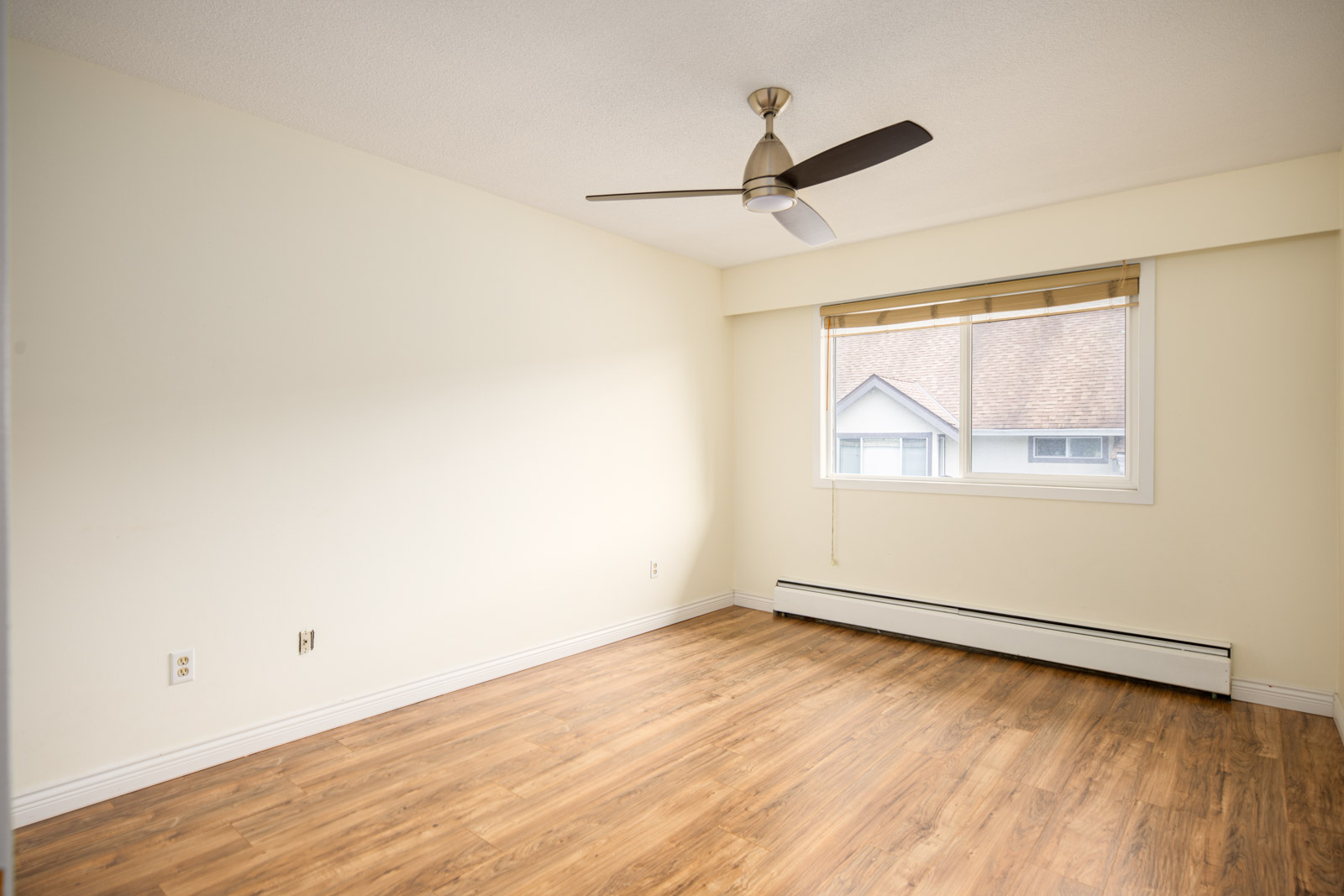 Empty room with light-colored walls, wood flooring, a window with blinds, ceiling fan, and baseboard heating.