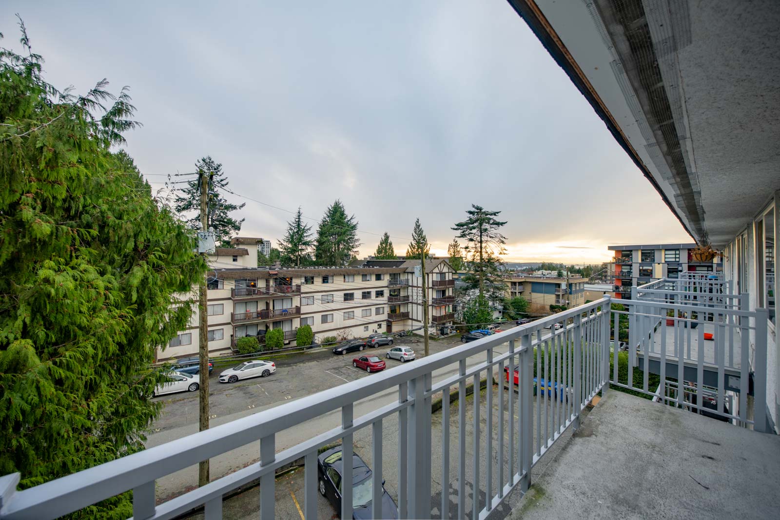 View from a balcony overlooking an apartment complex parking lot, with cars, trees, and neighboring buildings under a cloudy sky at sunset.