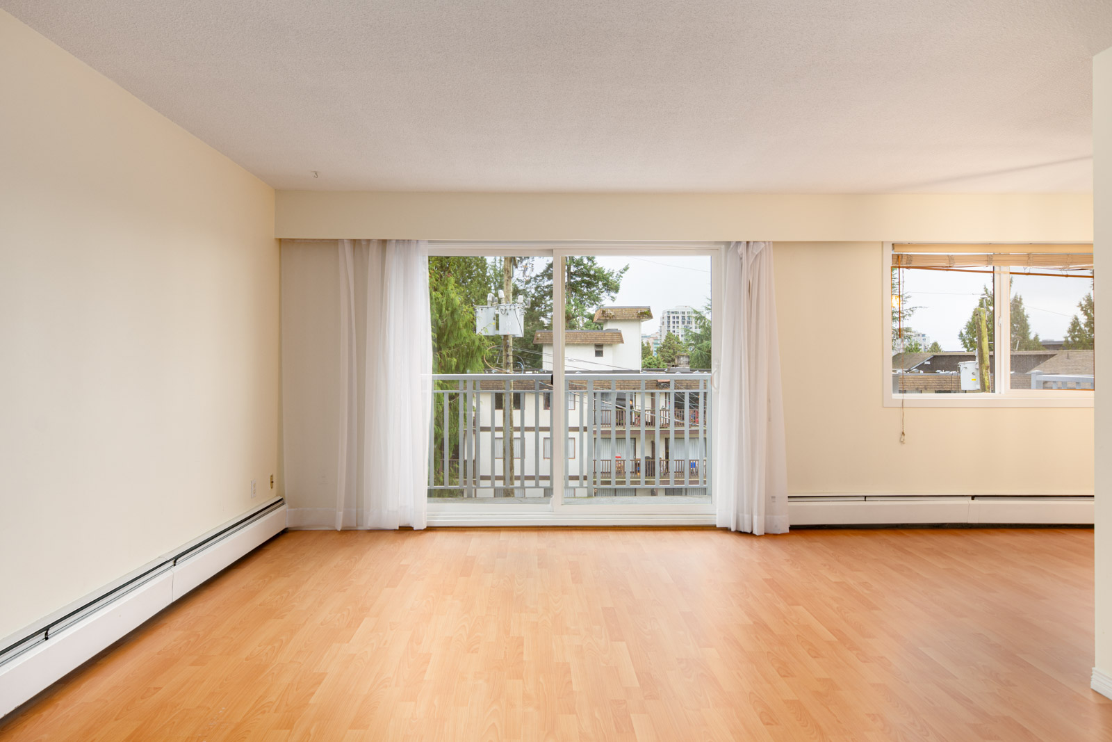 Empty room with light-colored walls, wood laminate flooring, a large window with sheer curtains, and a sliding glass door leading to a balcony with a view of trees and neighboring buildings.