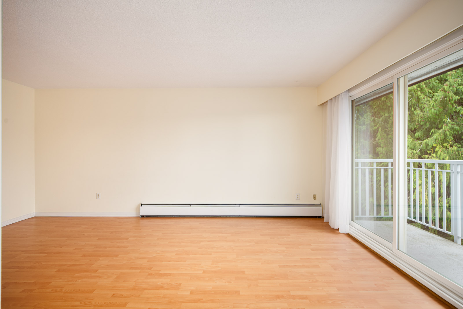 Empty room with light wood flooring, cream-colored walls, a baseboard heater, and large sliding glass doors leading to a balcony with a view of trees.