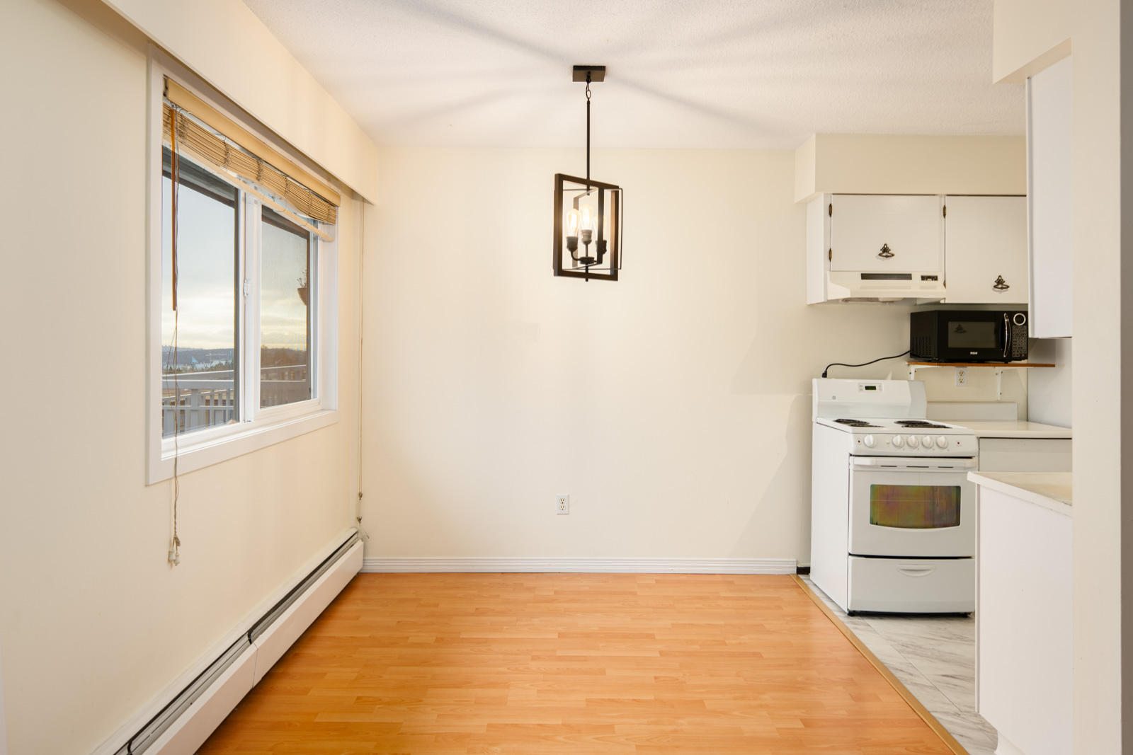 Small kitchen and dining area with light wood flooring, white cabinets, a stove, microwave, large window, and a modern ceiling light fixture.